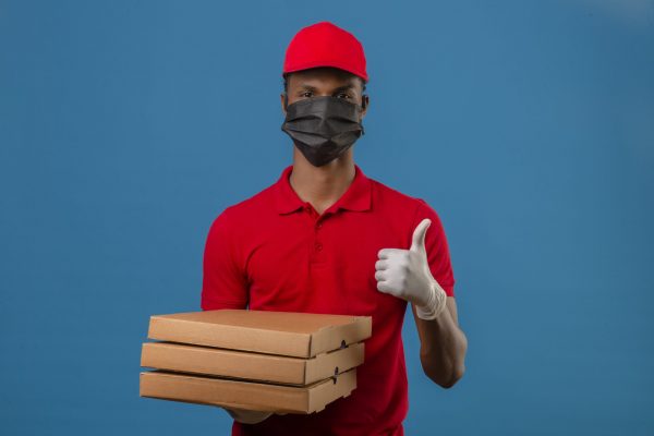 young-african-american-delivery-man-wearing-red-polo-shirt-and-cap-in-protective-mask-and-gloves-standing-with-stack-of-pizza-boxes-showing-thumbs-up-over-isolated-blue-min
